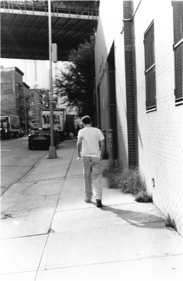 A young man in white waiting alone at a curb beneath scaffolding