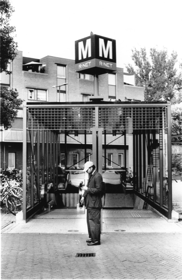 A man pausing at the mouth of a metro entrance, pigeons lined along the roof