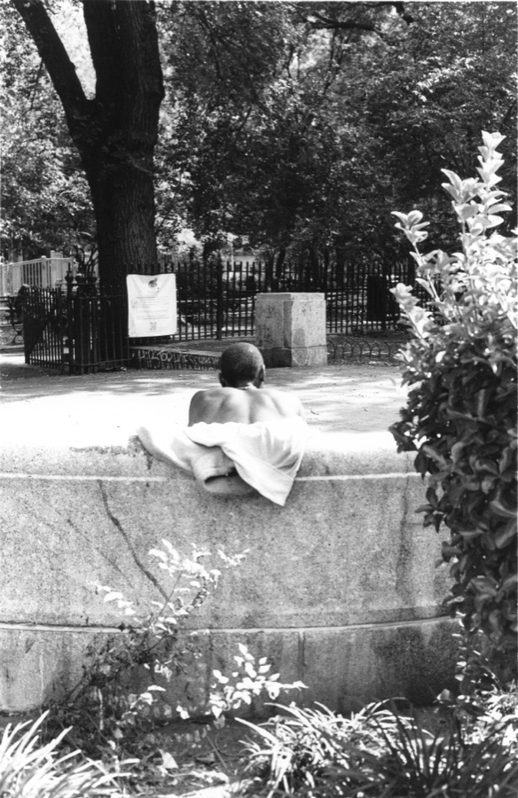 A shirtless man resting on a fountain rim in a fenced city park
