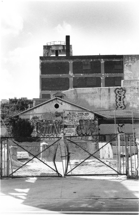 A chain-link gate padlocked across an empty lot, a graffiti-covered shed and derelict brick factory behind it