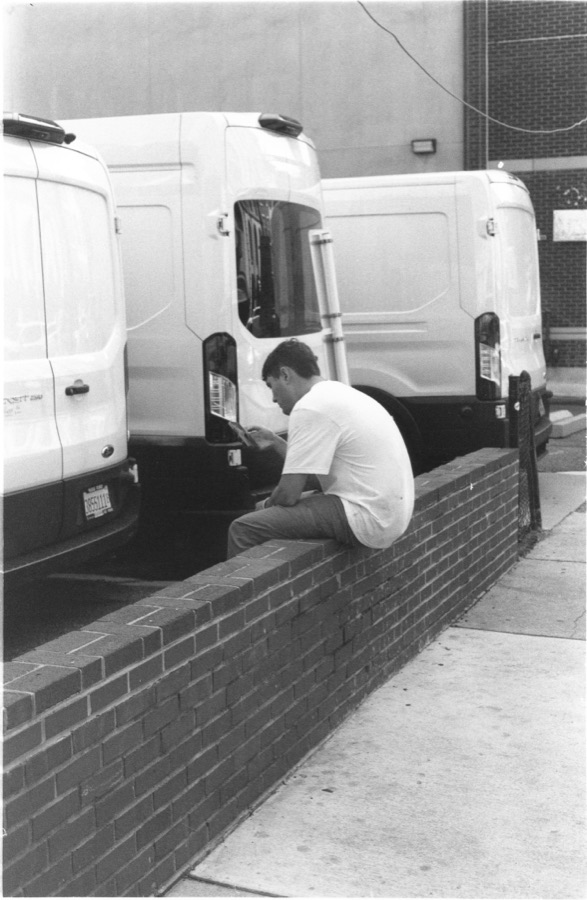 A man on a low brick wall absorbed in something in his hands, parked vans behind him