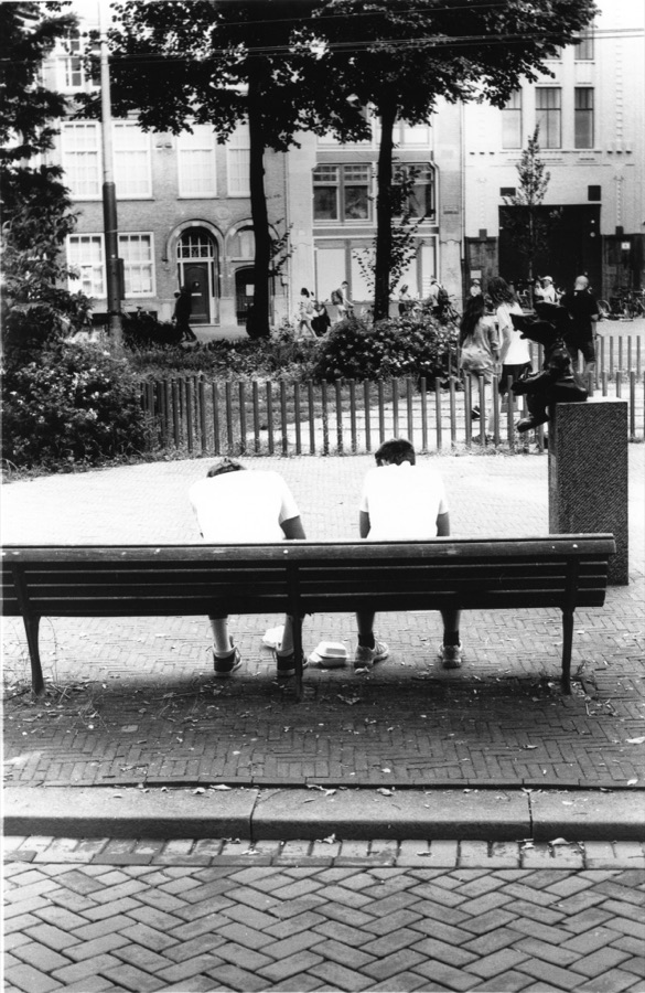 Two men sitting apart on a long bench, seen from behind
