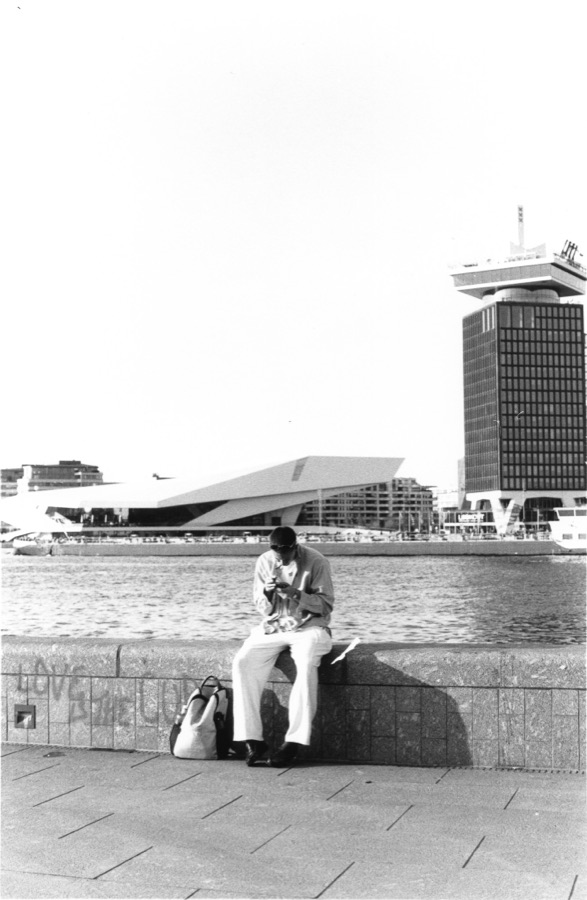 A man checking his phone on a wall above the water, a film museum and tower opposite