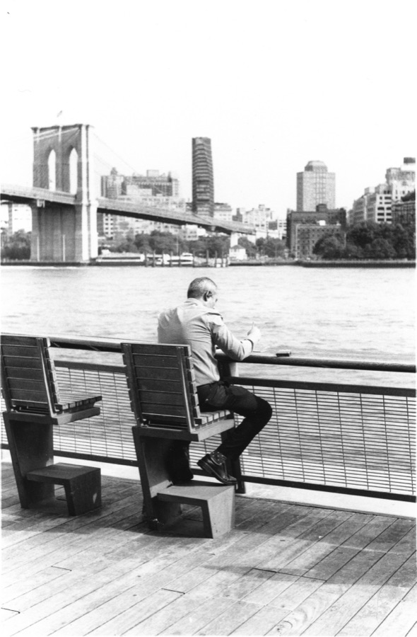 A man writing at the railing above the East River, the Brooklyn Bridge behind him