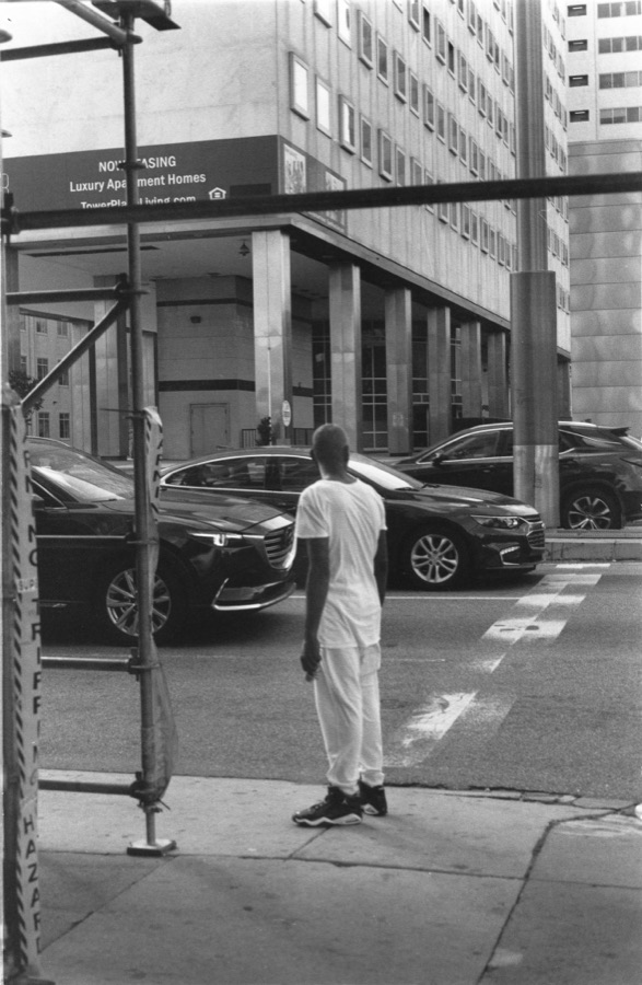 A young man in white waiting alone at a curb, scaffolding overhead and a Now Leasing banner across the street