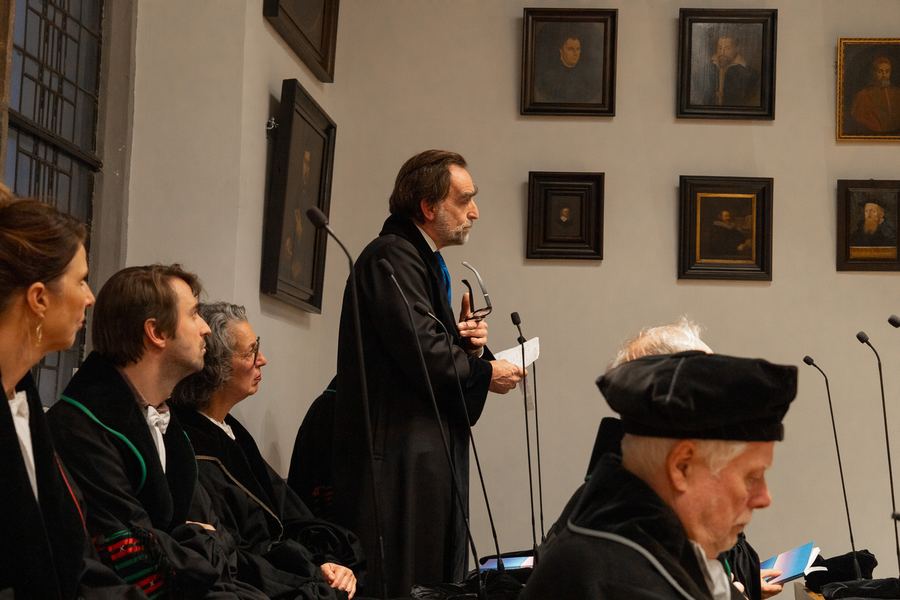 A professor in academic robes addressing the hall, the committee seated alongside