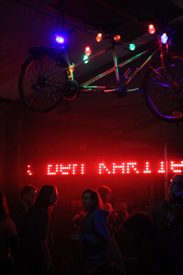 A tandem bicycle strung with coloured bulbs hanging above a crowd, a red LED sign glowing behind