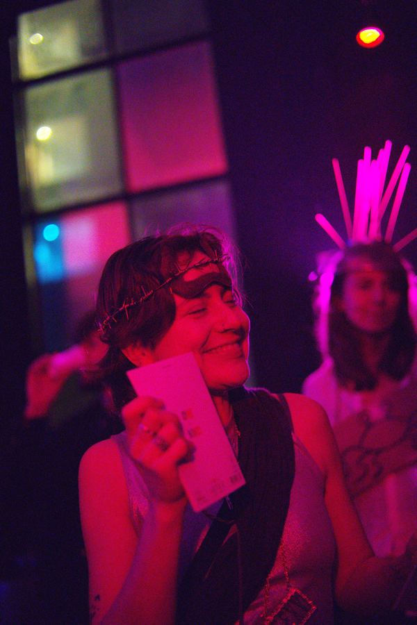 A guest in a wire crown and eye mask, smiling with a card in her hand under pink light